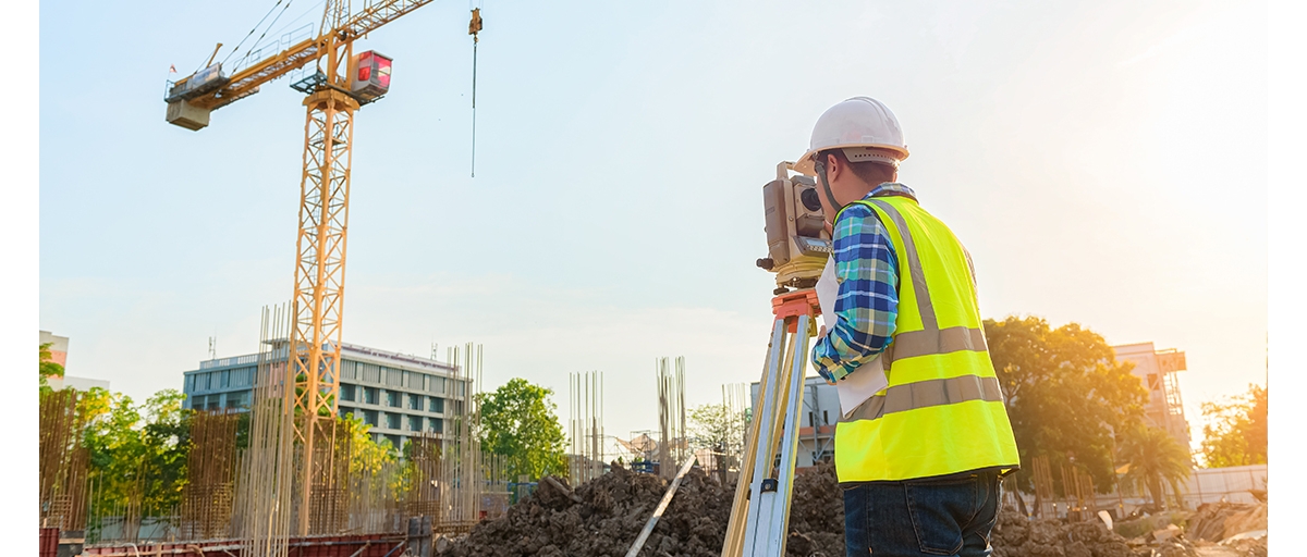 A person wearing a white helmet and reflective safety vest looking through a laser grade level to a construction site with a yellow crane.