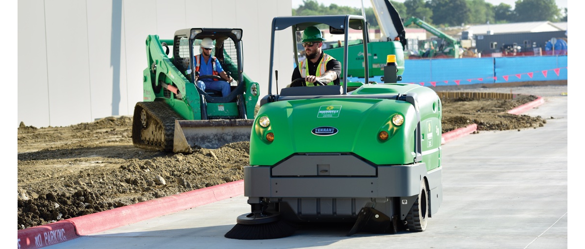 A man wearing a reflective safety vest and green helmet drives a pavement sweeper from Sunbelt Rentals, while another man uses an excavator to smooth a nearby dirt area.