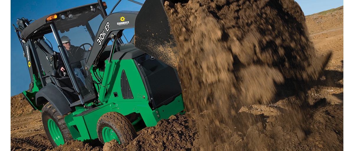 A person driving a green and black Sunbelt Rentals excavator dumps dirt from the excavator’s bucket.