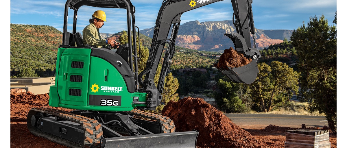 A man wearing a yellow construction helmet scooping dirt with a green Sunbelt Rentals excavator in front of a mountain vista.
