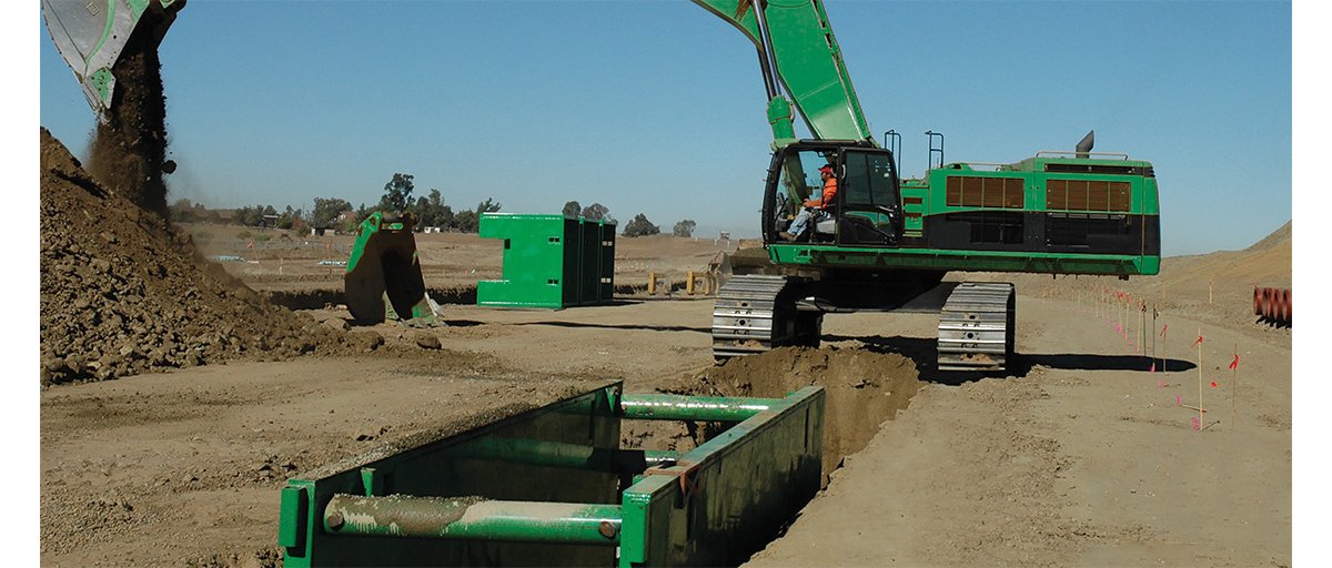 A construction worker uses a green excavator to dump dirt while digging a trench that is reinforced with green trench boxes.