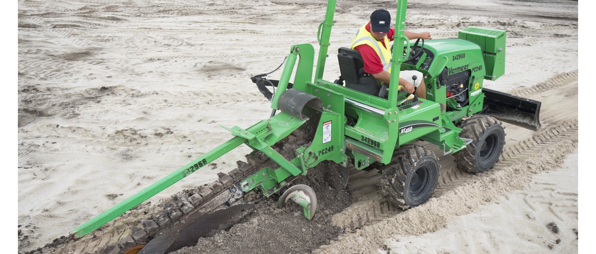 A man wearing a yellow safety vest over a red shirt digs a trench in sandy soil with a Sunbelt Rentals trencher.