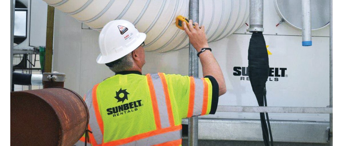A man wearing a white construction helmet and yellow safety vest with Sunbelt Rentals printed on the back uses a probe to monitor an HVAC system.
