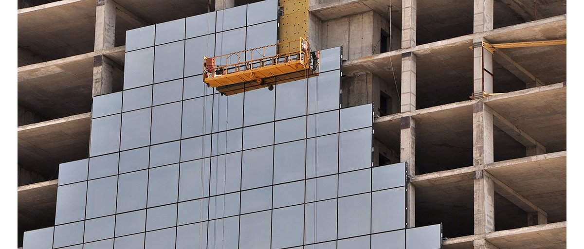 Construction workers use an orange elevated platform to install glass on the exterior of a high-rise building.
