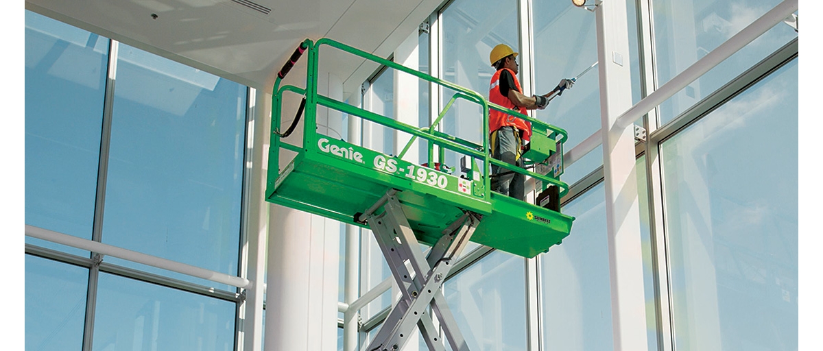 A construction worker wearing an orange safety vest and yellow helmet on a Sunbelt Rentals scissor lift cleaning a window.