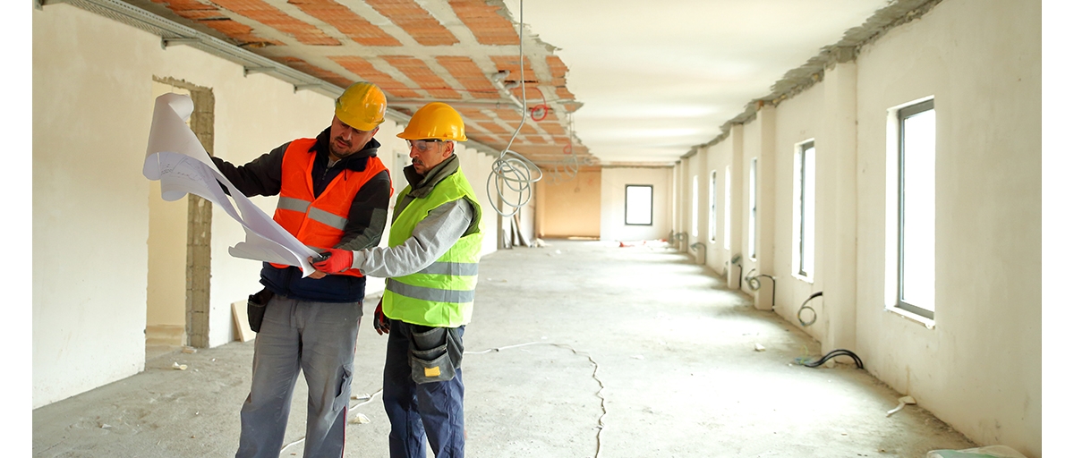 Two construction workers wearing reflective safety vests and yellow safety helmets look at a paper construction plan in a hallway of a building that’s under construction.