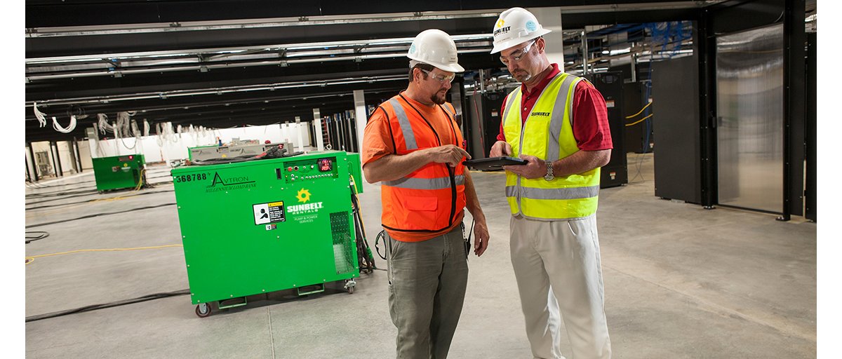 Two construction workers wearing white helmets and reflective safety vests looking at a tablet computer in front of a Sunbelt Rentals generator inside a partially finished building.