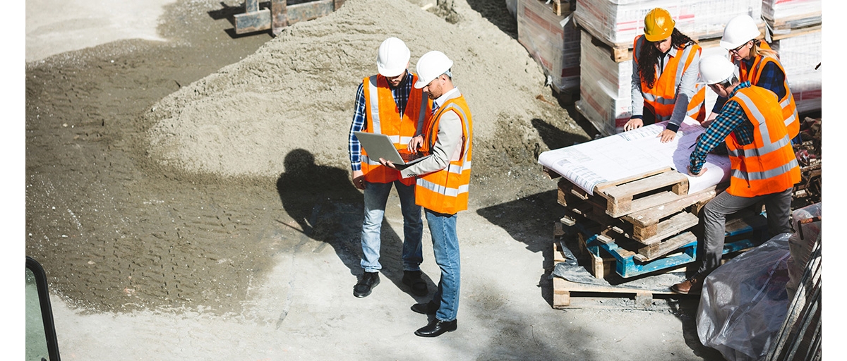 Two construction workers wearing orange safety vests and white helmets look at a laptop computer on a construction site, and nearby three construction workers look at printed plans laid out on a stack of wooden pallets.