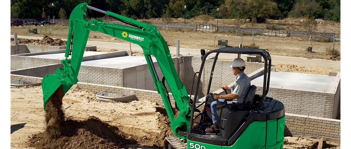 A construction worker wearing a white helmet uses a Sunbelt Rentals excavator to dig dirt.