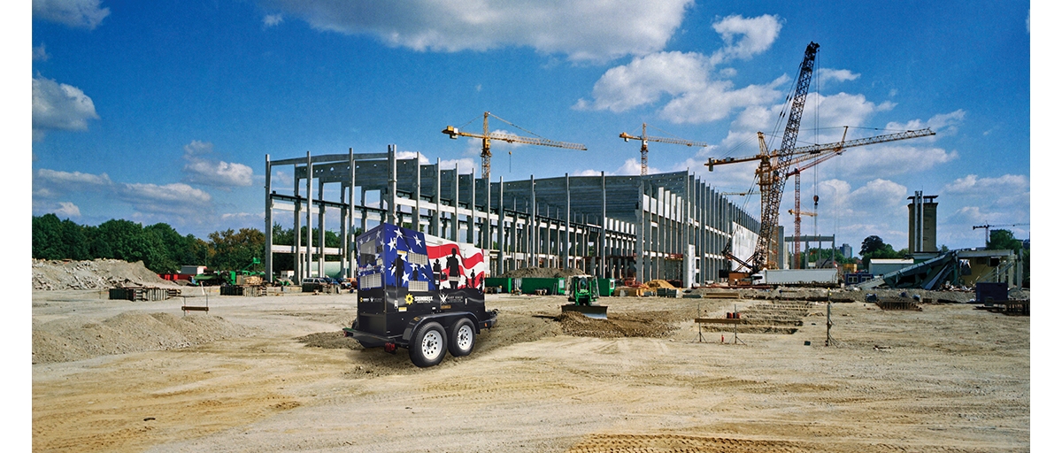 A generator with the American flag painted on it sits on a construction site where a metal-framed building is under construction.
