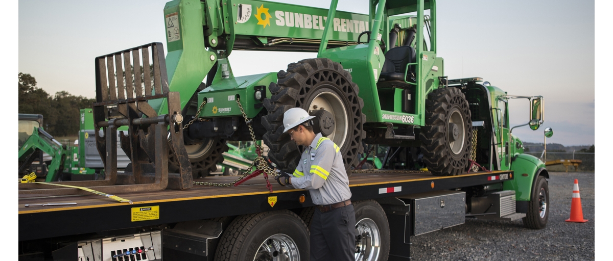 A man wearing a white helmet and gray reflective safety shirt secures a Telehandler Forklift from Sunbelt Rentals to a flatbed truck.