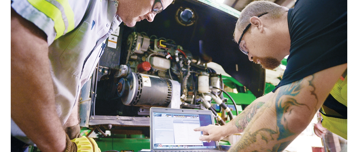 Two men wearing safety glasses look at a laptop computer next to an air compressor.