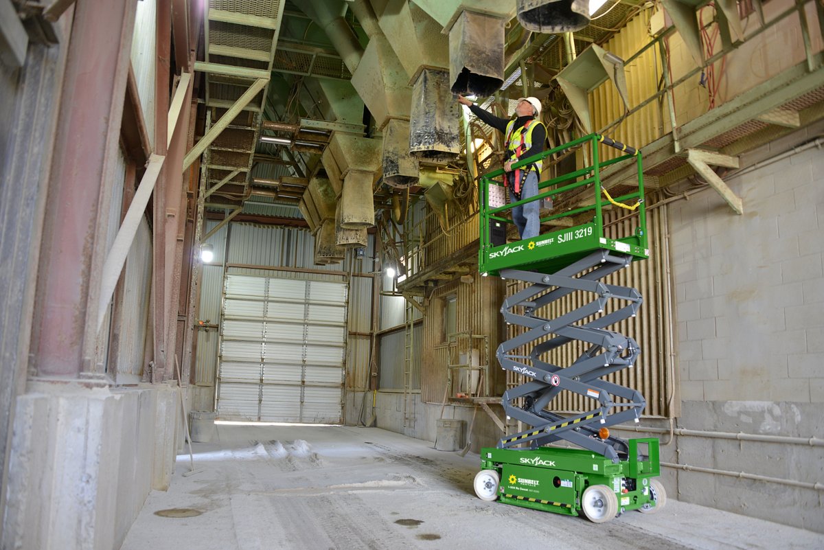 A person wearing a helmet and safety vest on a scissor lift in an empty warehouse.