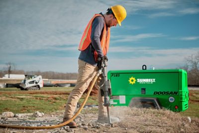 Someone using an air compressor to power a demolition hammer, at a construction site.