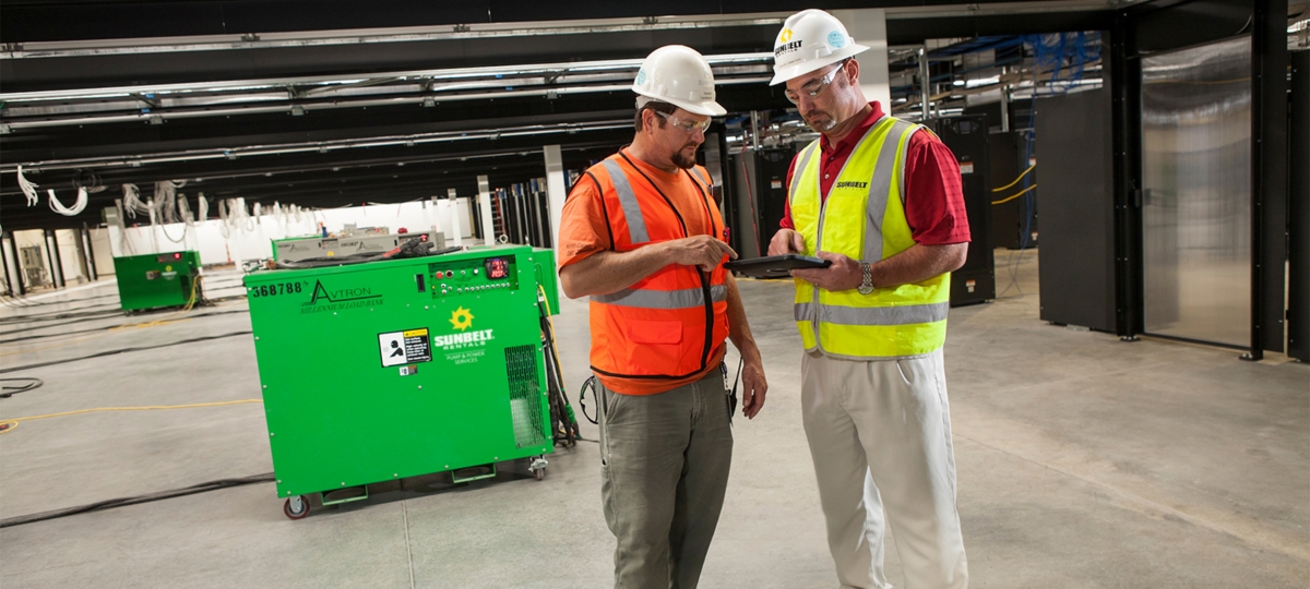 Two people standing together looking at a tablet device, in a building with load banks.
