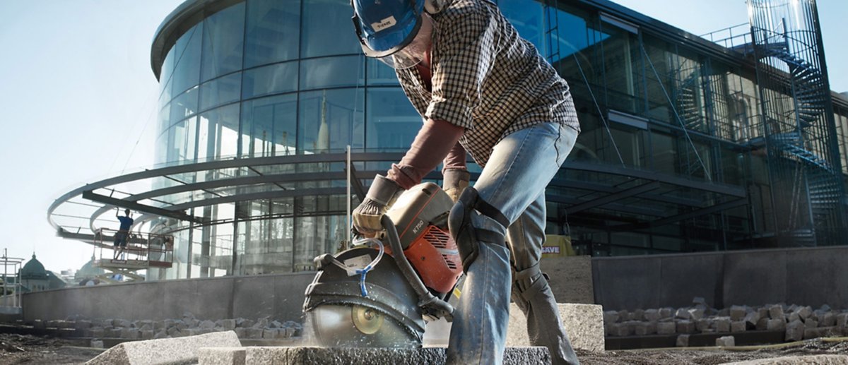 A man wearing a blue safety helmet uses a power tool to cut stone on the front of a large glass facade building.