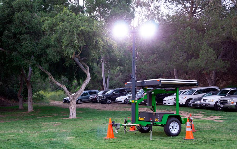 A light tower in the grass near parked cars.