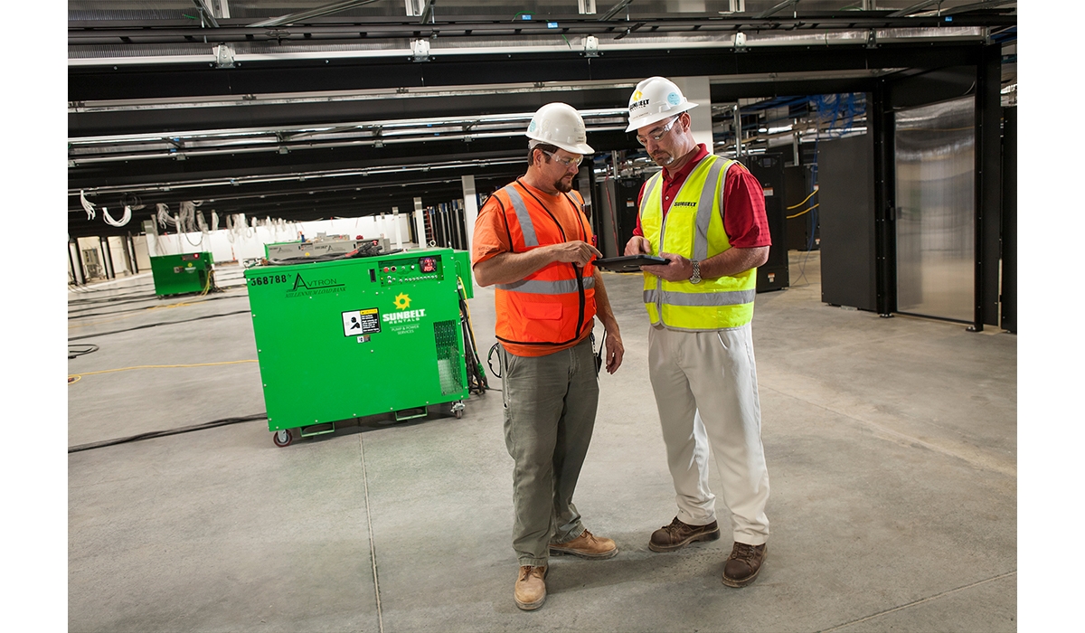 Two people standing together looking at a tablet device, in a building with load banks.