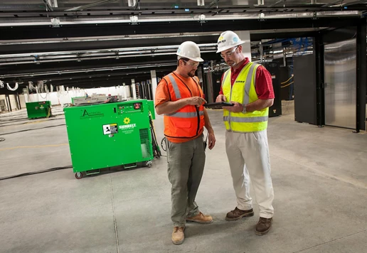Two people standing together looking at a tablet device, in a building with load banks.