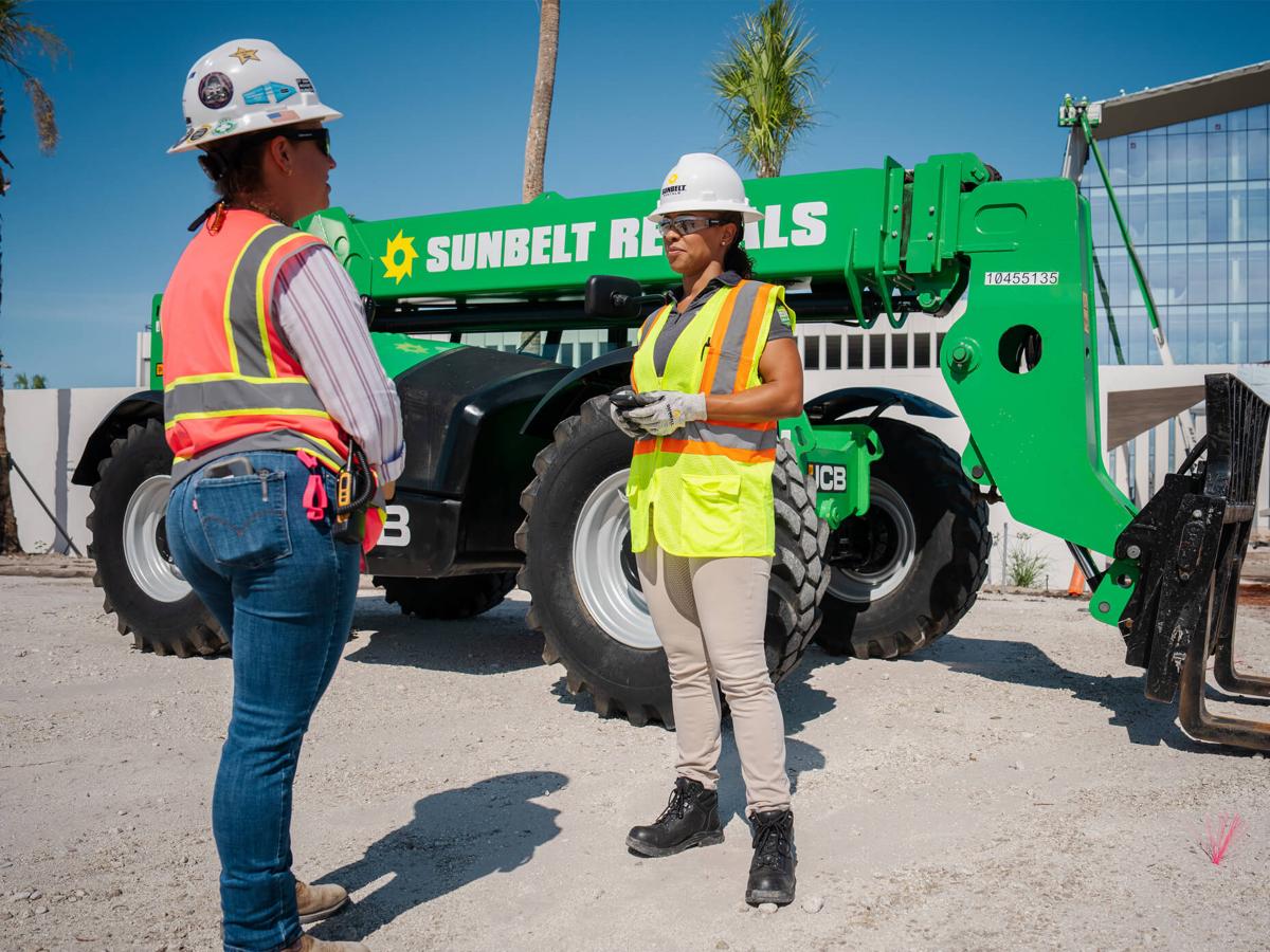 Two people wearing safety apparel stand next to a forklift from Sunbelt Rentals.