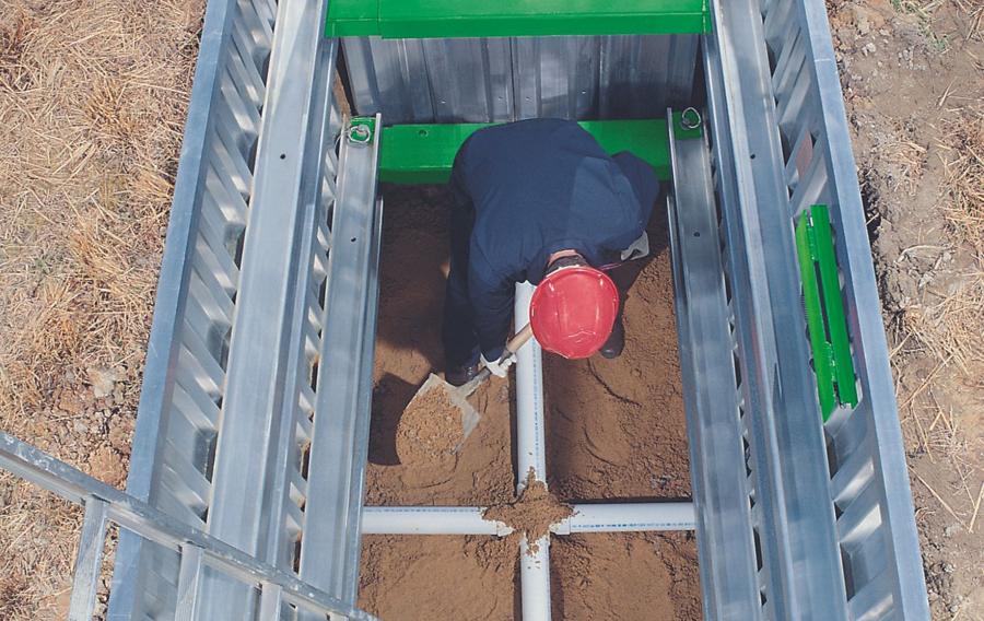 Someone wearing a helmet standing inside of a trench box, shoveling dirt.