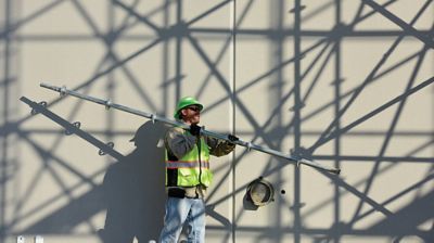A worker carrying scaffolding equipment in front an exterior wall.