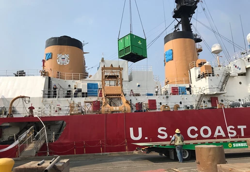 A U.S. Coast Guard ship docked at a port while a Sunbelt Rentals loading bank is lifted onto the ship.