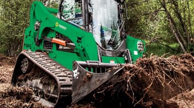 A skid steer picking up brush at a wooded job site.