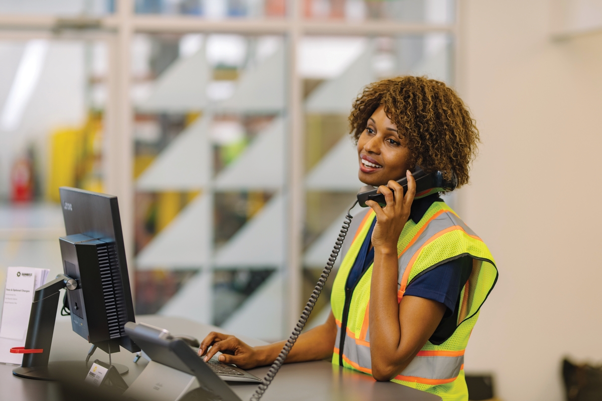 An employee wearing a safety vest stands at a desk and talks on the phone.